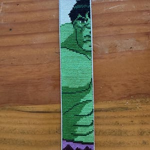 May include: A handmade bookmark featuring a cross-stitch design of the Hulk. The bookmark has a green and black Hulk image with purple accents and a white fringe at the top and bottom. The bookmark is approximately 8 inches long.