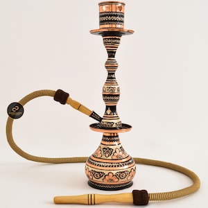 May include: A traditional copper hookah with intricate floral designs in black and white. The hookah has a tall, slender stem with a round base and a decorative bowl. The hookah comes with a hose and a mouthpiece.
