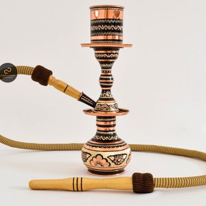 May include: A traditional copper hookah with intricate black and gold floral designs. The hookah has a round base, a tall stem, and a bowl on top. It comes with a hose and a mouthpiece.