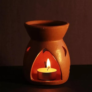 May include: A terracotta essential oil diffuser with a lit tea light candle inside. The diffuser has a rounded shape with cut-out designs and a small opening at the top. The warm glow of the candle illuminates the interior.