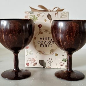 May include: Two dark brown coconut shell wine glasses with stems and round bases stand before a box with the text "vinty ceylon mart". The box is decorated with botanical illustrations and a ribbon.