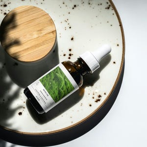 Puede incluir: Una botella de vidrio marrón con un gotero que contiene un líquido verde. La botella está etiquetada con "Moss Oil" y "100% Pure & Natural". La botella está sobre un plato de cerámica blanco con una tapa de madera.