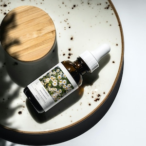 Op de afbeelding: Een bruine glazen fles met een witte druppelaar en een wit etiket met de tekst "Chamomile 100% Pure & Natural" en een afbeelding van kamillebloemen. De fles staat op een wit keramiek bord met bruine vlekken.