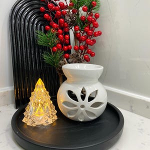 Puede incluir: Difusor de aceite de cerámica blanca con un diseño floral, un árbol de Navidad de vidrio transparente con luces blancas cálidas y una bandeja negra con una rama de bayas rojas.