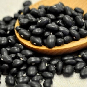 Puede incluir: Primer plano de frijoles negros en una cuchara de madera. Los frijoles están esparcidos alrededor de la cuchara sobre una superficie marrón claro.