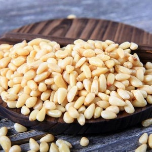 May include: A close-up of a wooden plate filled with pine nuts. The nuts are light brown and have a slightly oval shape. The plate is sitting on a wooden surface.