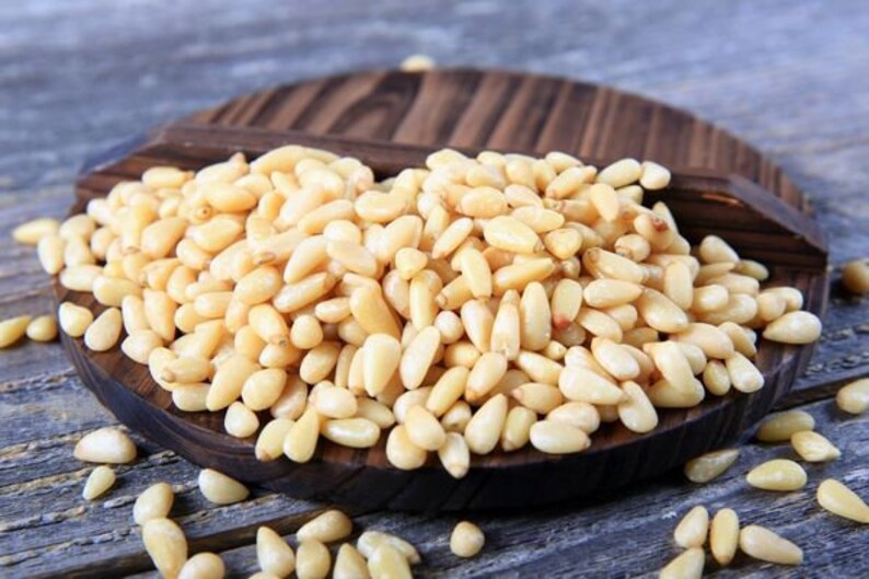 May include: A close-up of a wooden plate filled with pine nuts. The nuts are light brown and have a slightly oval shape. The plate is sitting on a wooden surface.
