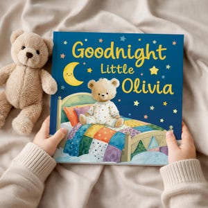 May include: A children's book titled "Goodnight Little Olivia" with a teddy bear illustration on the cover. The cover is dark blue with a crescent moon, stars, and the book's title in yellow. A stuffed teddy bear is next to the book.