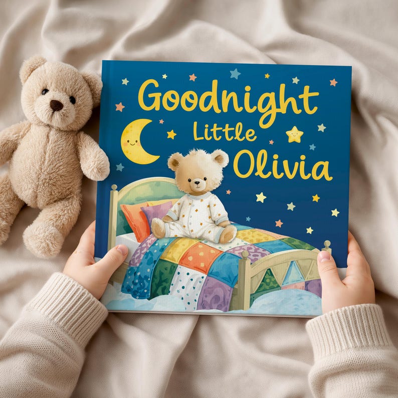May include: A children's book titled "Goodnight Little Olivia" with a teddy bear illustration on the cover. The cover is dark blue with a crescent moon, stars, and the book's title in yellow. A stuffed teddy bear is next to the book.
