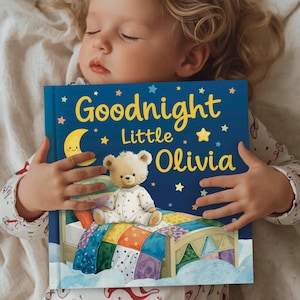 May include: A child asleep in bed holds a blue children's book titled "Goodnight Little Olivia." The book cover features a teddy bear in pajamas sitting on a bed, with a crescent moon and stars. The child is wearing pajamas.