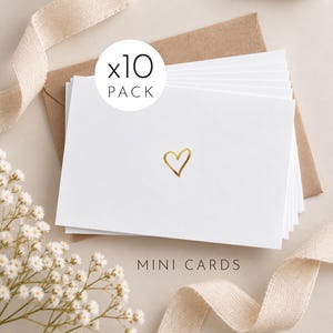 May include: A pack of ten white mini cards featuring a small gold foil heart in the centre. The cards are stacked on top of a brown envelope. The text x10 PACK and MINI CARDS is printed on the image. A light beige ribbon and white flowers are visible.