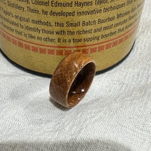 Puede incluir: Un anillo de madera hecho a mano con un patr&oacute;n de vetas de madera natural. El anillo es de color marr&oacute;n c&aacute;lido y parece estar hecho de un barril. El anillo est&aacute; sobre una superficie blanca.
