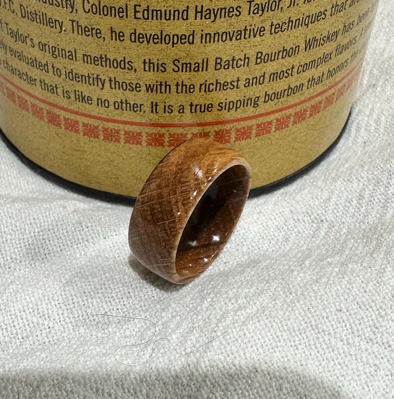 Puede incluir: Un anillo de madera hecho a mano con un patr&oacute;n de vetas de madera natural. El anillo es de color marr&oacute;n c&aacute;lido y parece estar hecho de un barril. El anillo est&aacute; sobre una superficie blanca.