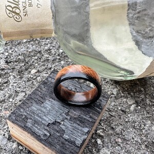May include: A handcrafted wooden ring with a dark, glossy finish on the outer band and a lighter, natural wood grain on the top. The ring is displayed on a piece of charred wood, with a blurred bottle in the background.