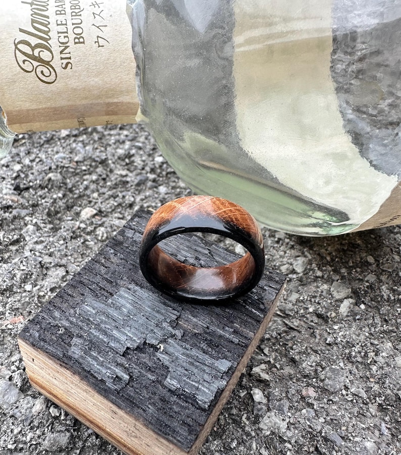 May include: A handcrafted wooden ring with a dark, glossy finish on the outer band and a lighter, natural wood grain on the top. The ring is displayed on a piece of charred wood, with a blurred bottle in the background.