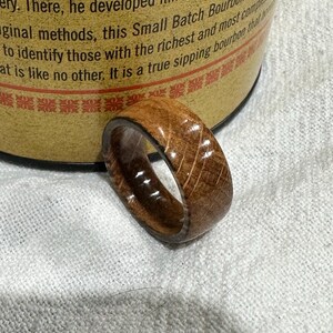 Puede incluir: Un anillo de madera con un interior marr&oacute;n oscuro y un exterior marr&oacute;n claro. El anillo tiene una superficie texturizada con patrones de vetas de madera visibles. El anillo es probablemente hecho a mano y es una joya &uacute;nica.