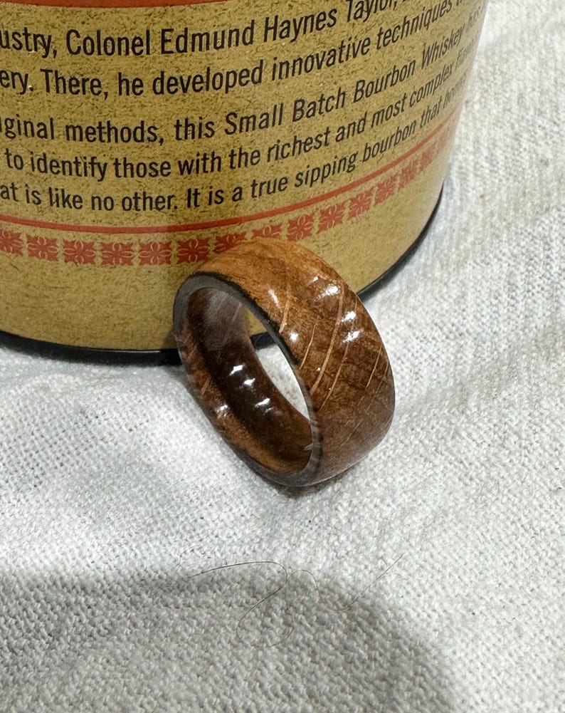 Puede incluir: Un anillo de madera con un interior marr&oacute;n oscuro y un exterior marr&oacute;n claro. El anillo tiene una superficie texturizada con patrones de vetas de madera visibles. El anillo es probablemente hecho a mano y es una joya &uacute;nica.
