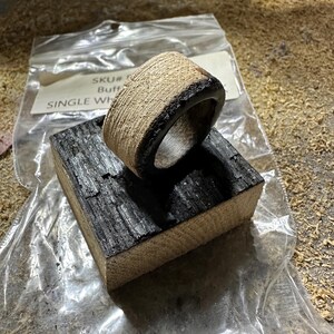 May include: A handcrafted wooden ring with a textured, light-colored exterior and a dark, charred interior and edge. The ring rests on a small, charred wooden block. The ring is in a clear plastic bag.