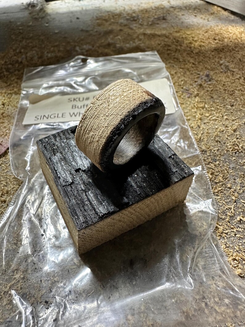 May include: A handcrafted wooden ring with a textured, light-colored exterior and a dark, charred interior and edge. The ring rests on a small, charred wooden block. The ring is in a clear plastic bag.