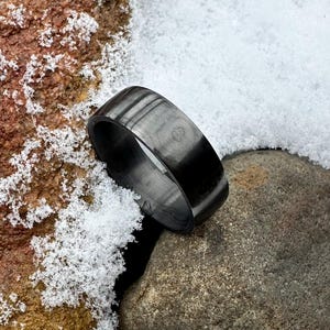 以下が含まれることがあります： 滑らかで磨かれた仕上げの黒いカーボンファイバーリング。リングは雪で覆われた岩の上に置かれています。