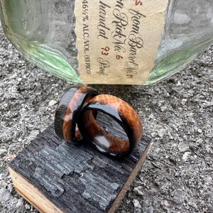 May include: Two wooden rings with a dark wood and light wood grain design, resting on a piece of charred wood. The rings are near a bottle with text that reads "from Barrel No. 6".