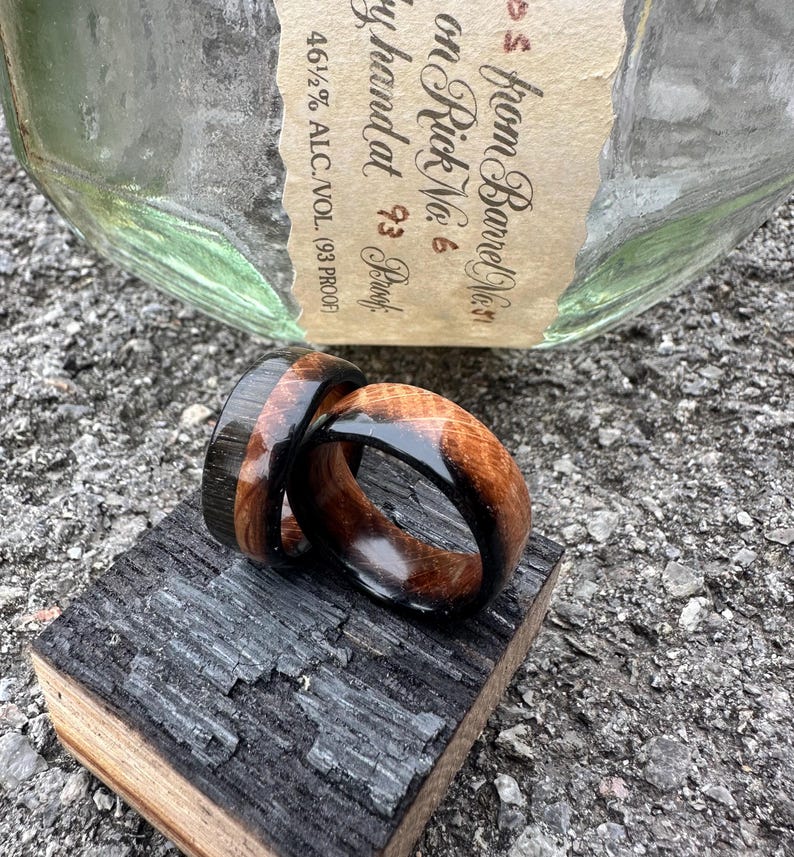 May include: Two wooden rings with a dark wood and light wood grain design, resting on a piece of charred wood. The rings are near a bottle with text that reads "from Barrel No. 6".