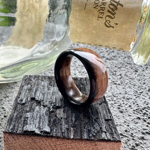 May include: A wooden ring with a dark, glossy finish on the inner band and a lighter, natural wood grain on the outer surface. The ring is displayed on a piece of dark, textured wood.