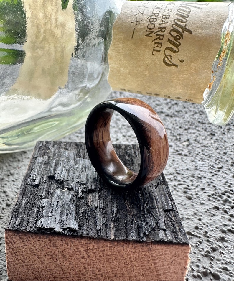 May include: A wooden ring with a dark, glossy finish on the inner band and a lighter, natural wood grain on the outer surface. The ring is displayed on a piece of dark, textured wood.