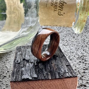 May include: A wooden ring with a polished finish, showcasing the natural grain of the wood. The ring is set against a backdrop of a charred wood block and a partially visible glass bottle. The ring's warm tones contrast with the darker wood.