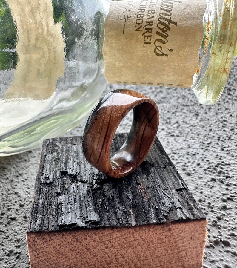 May include: A wooden ring with a polished finish, showcasing the natural grain of the wood. The ring is set against a backdrop of a charred wood block and a partially visible glass bottle. The ring's warm tones contrast with the darker wood.