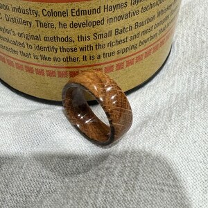 Puede incluir: Un anillo de madera con un patr&oacute;n de vetas de madera natural. El anillo es de color marr&oacute;n medio y tiene un acabado liso y pulido. El anillo est&aacute; sobre una superficie blanca.