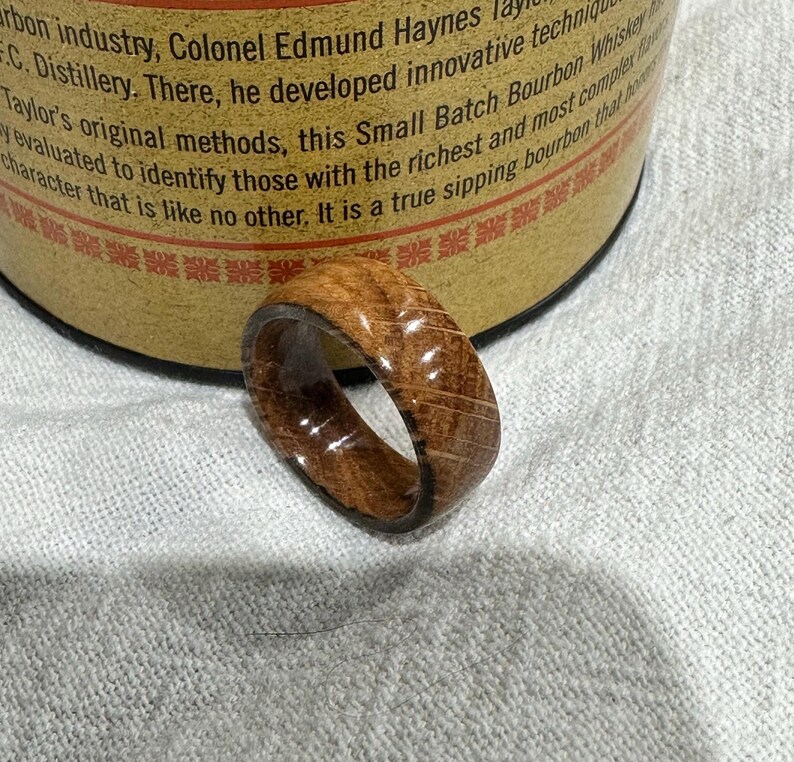 Puede incluir: Un anillo de madera con un patr&oacute;n de vetas de madera natural. El anillo es de color marr&oacute;n medio y tiene un acabado liso y pulido. El anillo est&aacute; sobre una superficie blanca.
