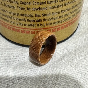 Puede incluir: Un anillo de madera de color marr&oacute;n claro con vetas visibles. El anillo se encuentra frente a un barril de whisky, lo que sugiere una conexi&oacute;n con la industria de las bebidas espirituosas. El dise&ntilde;o del anillo es simple y elegante.