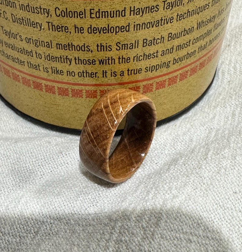 Puede incluir: Un anillo de madera de color marr&oacute;n claro con vetas visibles. El anillo se encuentra frente a un barril de whisky, lo que sugiere una conexi&oacute;n con la industria de las bebidas espirituosas. El dise&ntilde;o del anillo es simple y elegante.