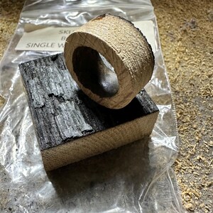 May include: A wooden ring and a rectangular block, both with a charred, dark gray surface and a light brown, textured interior. The ring sits atop the block, both appearing to be freshly cut. The items are on a surface covered in wood shavings.