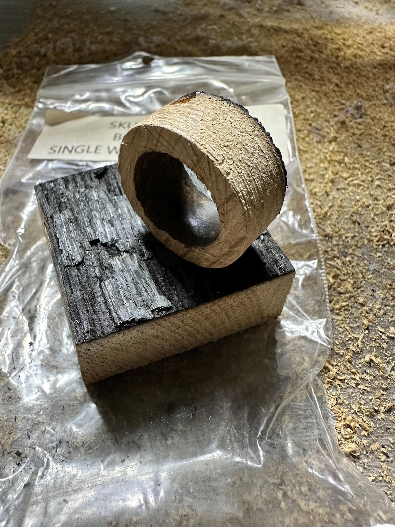 May include: A wooden ring and a rectangular block, both with a charred, dark gray surface and a light brown, textured interior. The ring sits atop the block, both appearing to be freshly cut. The items are on a surface covered in wood shavings.