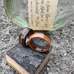 May include: Two wooden rings, one with a dark stripe, rest on a charred wood block. The rings are made of light brown wood with visible grain. A bottle with a label that reads "from Barrel No. 31" is in the background.
