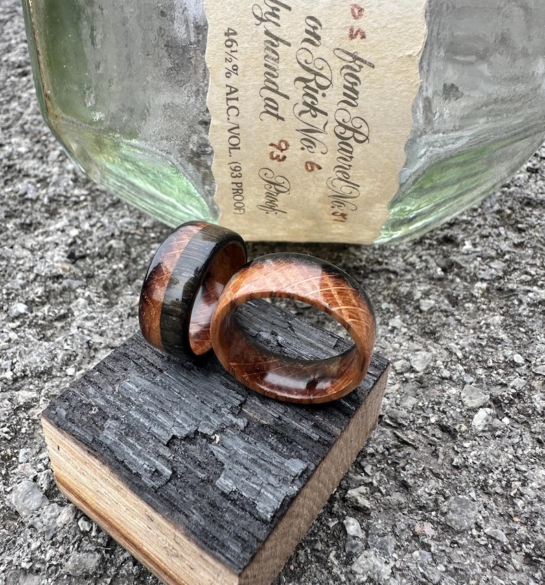 May include: Two wooden rings, one with a dark stripe, rest on a charred wood block. The rings are made of light brown wood with visible grain. A bottle with a label that reads "from Barrel No. 31" is in the background.