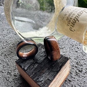 May include: Two wooden rings with a dark brown and black design are displayed on a piece of charred wood. The rings have a polished finish, showcasing the wood grain. A clear glass bottle is in the background.