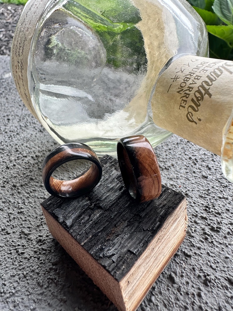 May include: Two wooden rings with a dark brown and black design are displayed on a piece of charred wood. The rings have a polished finish, showcasing the wood grain. A clear glass bottle is in the background.