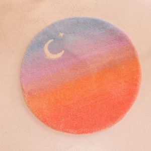 May include: Round rug with a gradient design in shades of blue, pink, and orange, resembling a sunset. A white crescent moon and star are featured in the blue section. The rug is a soft, plush material.