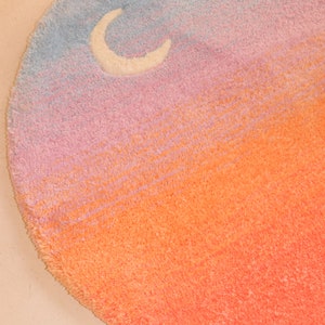May include: A round, plush rug with a gradient design transitioning from blue to pink to orange. A white crescent moon and star are in the blue section. The rug is a decorative home accent.
