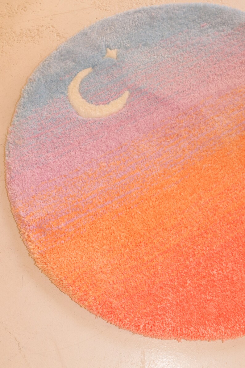 May include: A round, plush rug with a gradient design transitioning from blue to pink to orange. A white crescent moon and star are in the blue section. The rug is a decorative home accent.