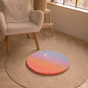 May include: A round, hand-tufted rug with a gradient design in shades of orange, pink, blue, and purple. A white crescent moon and star are featured on the top. The rug is placed on a larger, natural fiber jute rug.