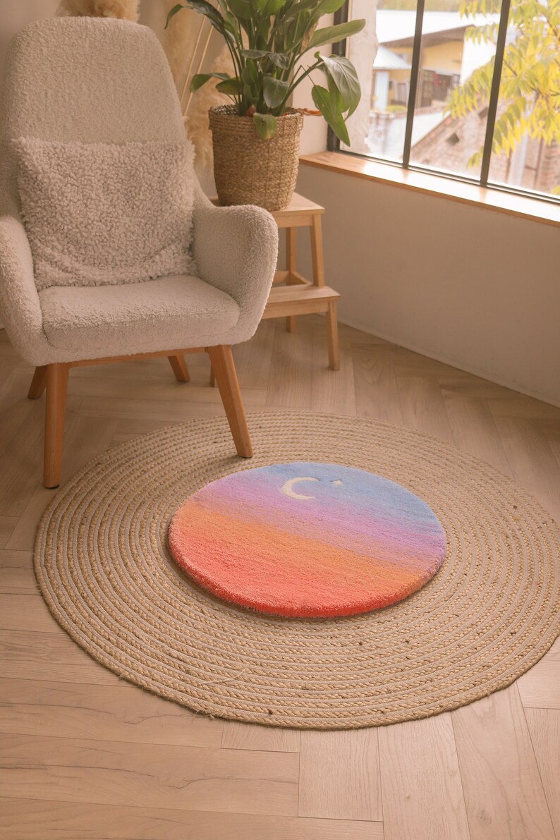 May include: A round, hand-tufted rug with a gradient design in shades of orange, pink, blue, and purple. A white crescent moon and star are featured on the top. The rug is placed on a larger, natural fiber jute rug.