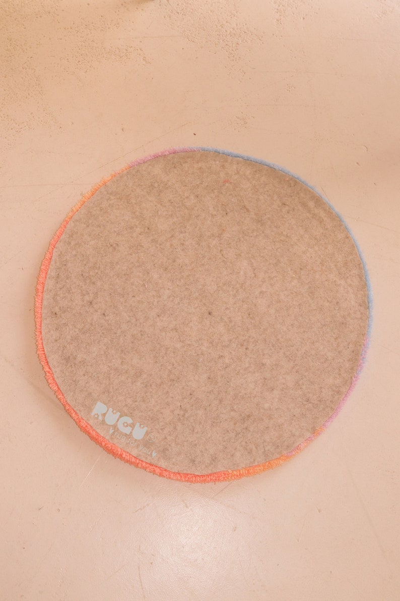 May include: A round rug with a light brown center and a colorful border. The border transitions from orange to pink to blue. The rug has the text "RUGU" in the lower left corner.