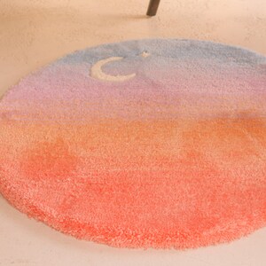 May include: A round, plush rug with a gradient design. The colors transition from a vibrant coral at the bottom to orange, pink, and finally, a light blue at the top. A white crescent moon and star are featured.