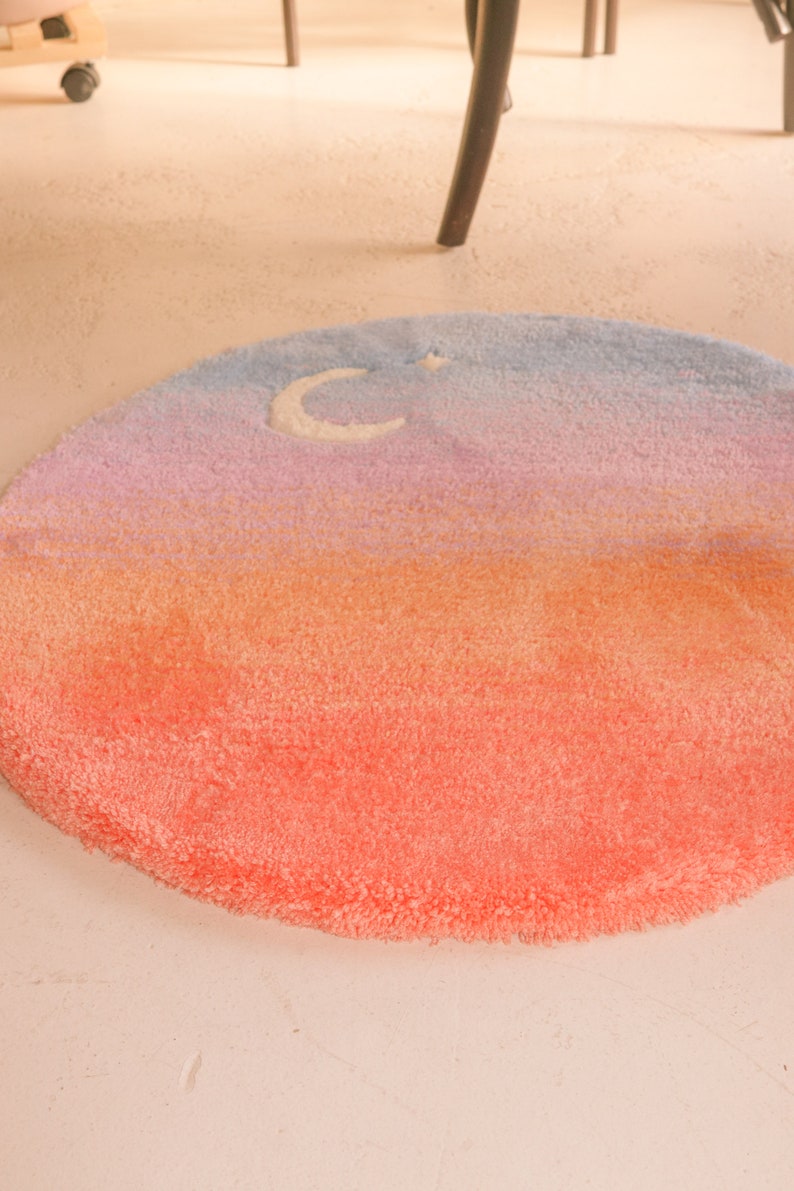 May include: A round, plush rug with a gradient design. The colors transition from a vibrant coral at the bottom to orange, pink, and finally, a light blue at the top. A white crescent moon and star are featured.
