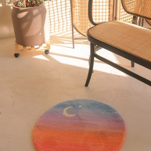 May include: A round rug with a gradient design, transitioning from orange to pink, purple, and blue. A white crescent moon and star are embroidered on the blue section. The rug is on a white floor.