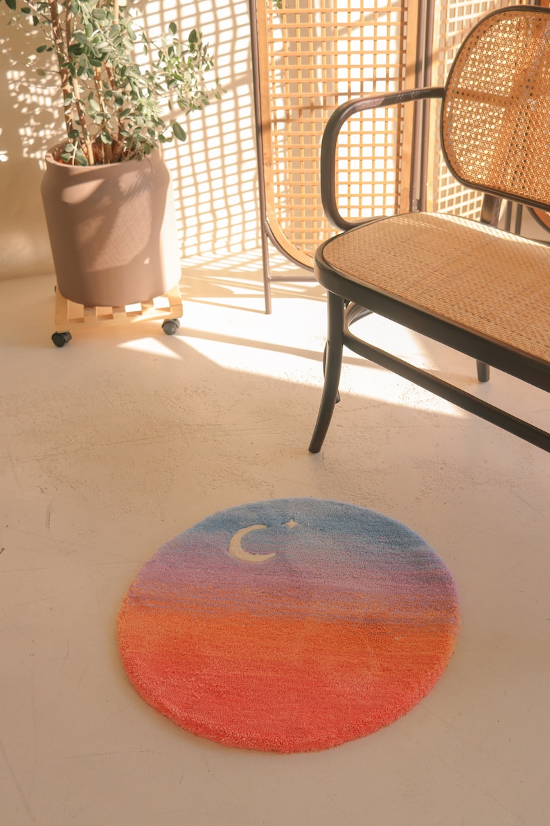 May include: A round rug with a gradient design, transitioning from orange to pink, purple, and blue. A white crescent moon and star are embroidered on the blue section. The rug is on a white floor.